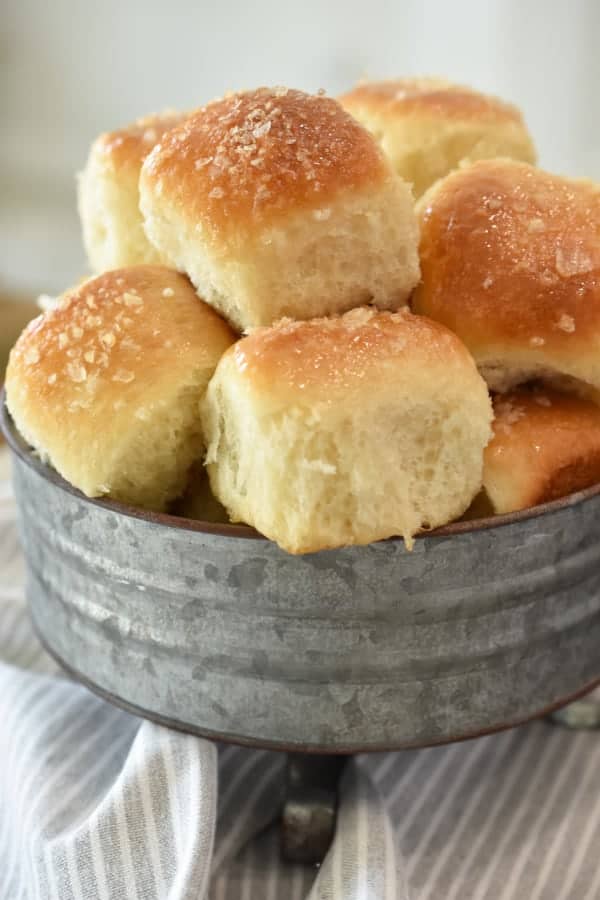 Steakhouse Dinner Rolls piled high and ready to eat. They are in a decorative tin container and a grey and white cloth is draped around it.