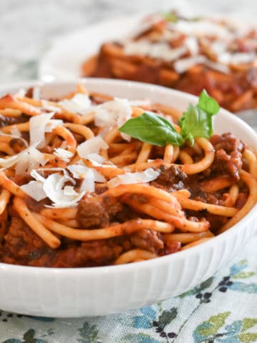 Bucatini pasta is mixed with Italian Meat Sauce and piled into a white bowl. Ribbons of Parmesan cheese and a sprig of basil garnish the top of the pasta with sauce.