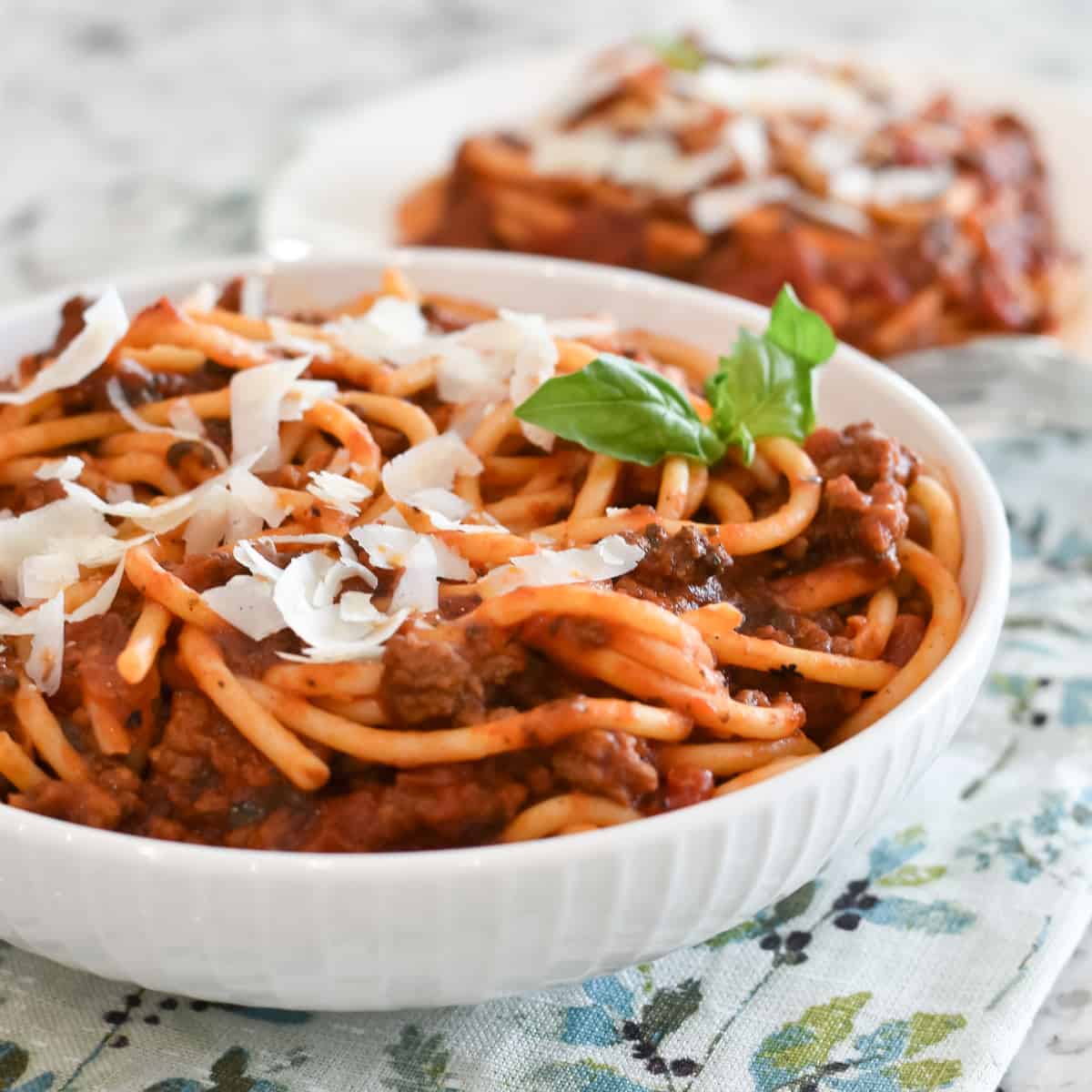 Bucatini pasta is mixed with rich meat sauce and piled into a white bowl. Ribbons of Parmesan cheese and a sprig of basil garnish the top of the pasta with sauce.