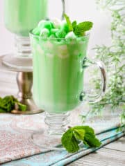 A light green mint-flavored hot chocolate in a tall glass mug is garnished with mini marshmallows and a drizzle of crème de menthe cordial.