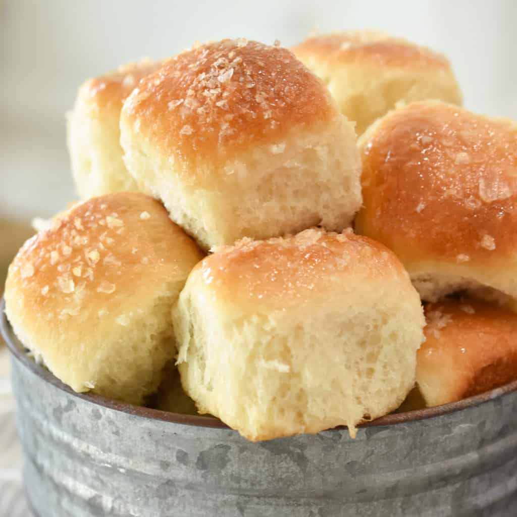 Dinner rolls glistening with butter and honey, are generously sprinkled with flaked sea salt and piled high in a rustic serving container.
