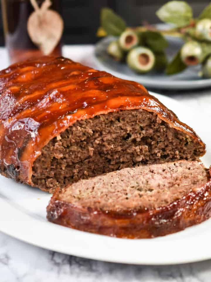 Oval shaped meatloaf covered with bacon and barbecue sauce on white serving platter with fall decoration.