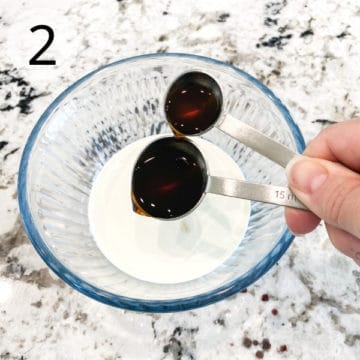 Dark colored Irish Cream syrup is being added to heavy cream in a small blue-tinted clear glass bowl.