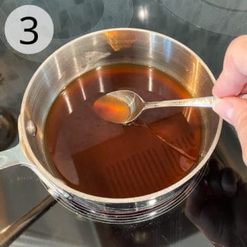 Brown sugar is dissolving in the warmed water in a small saucepan. A hand holds a teaspoon to stir the mixture.