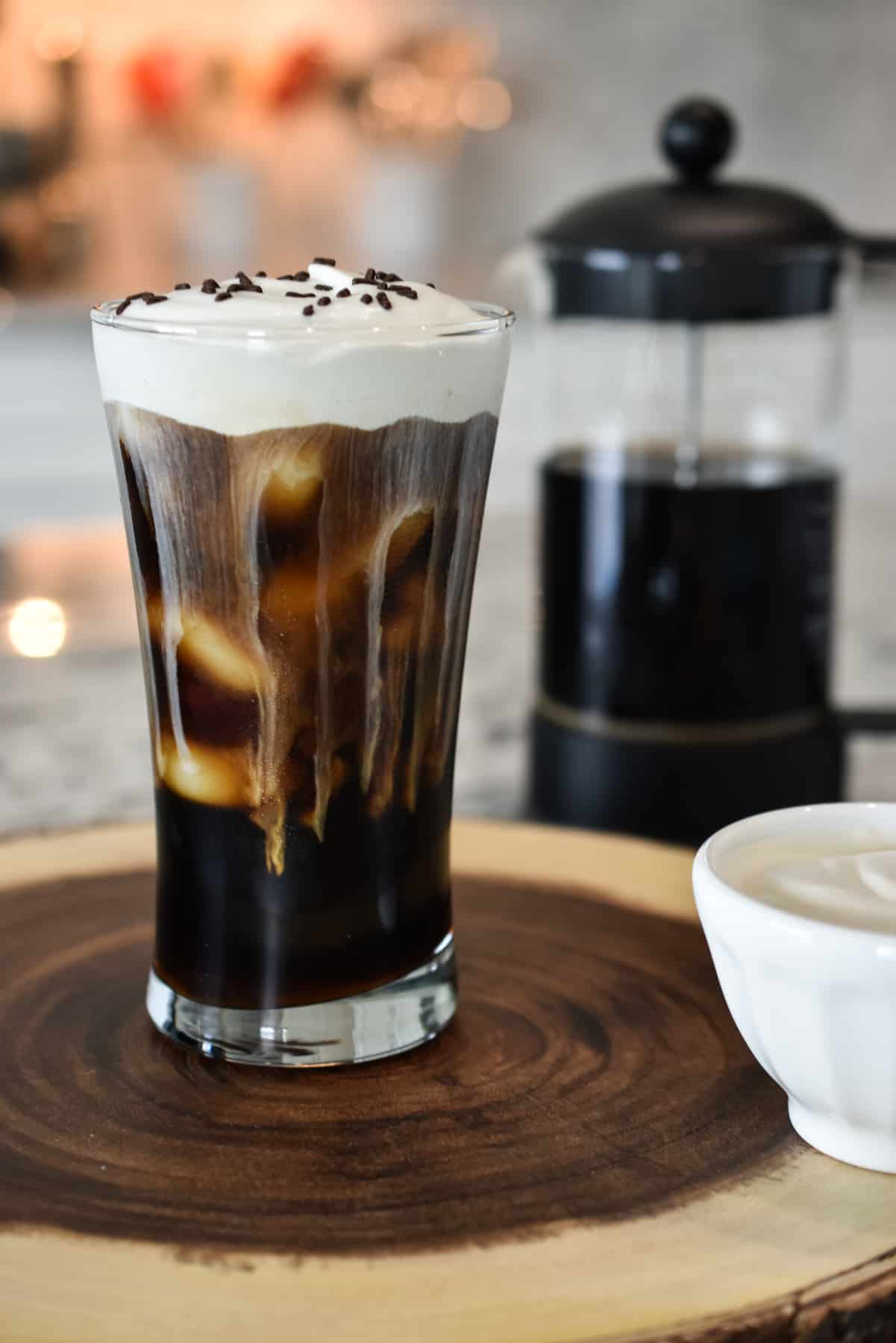 Portrait view of a tall, iced, black coffee is sweetened with dark brown sugar and topped with fluffy Irish cream flavored whipped cream.