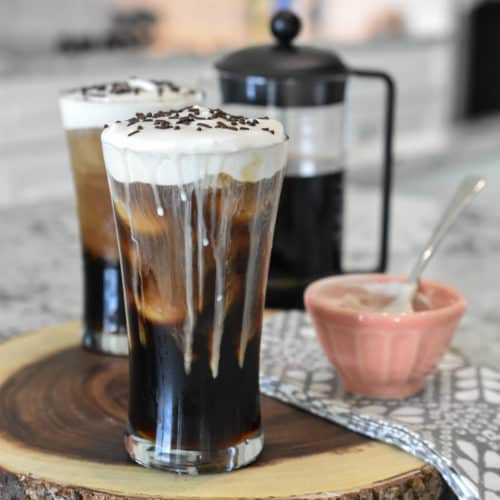 Square image of two iced coffees that are topped with whipped cream. A half-filled French press coffee maker is in the background. A peach bowl with traces of whipped cream is in the foreground.