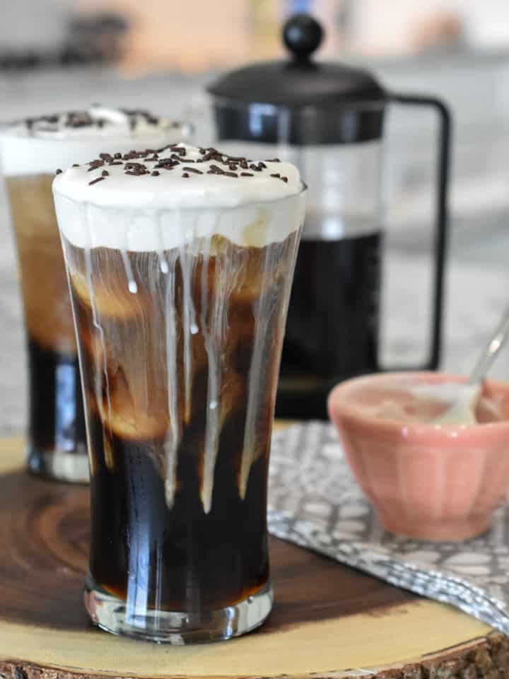 Square image of two iced coffees that are topped with whipped cream. A half-filled French press coffee maker is in the background. A peach bowl with traces of whipped cream is in the foreground.
