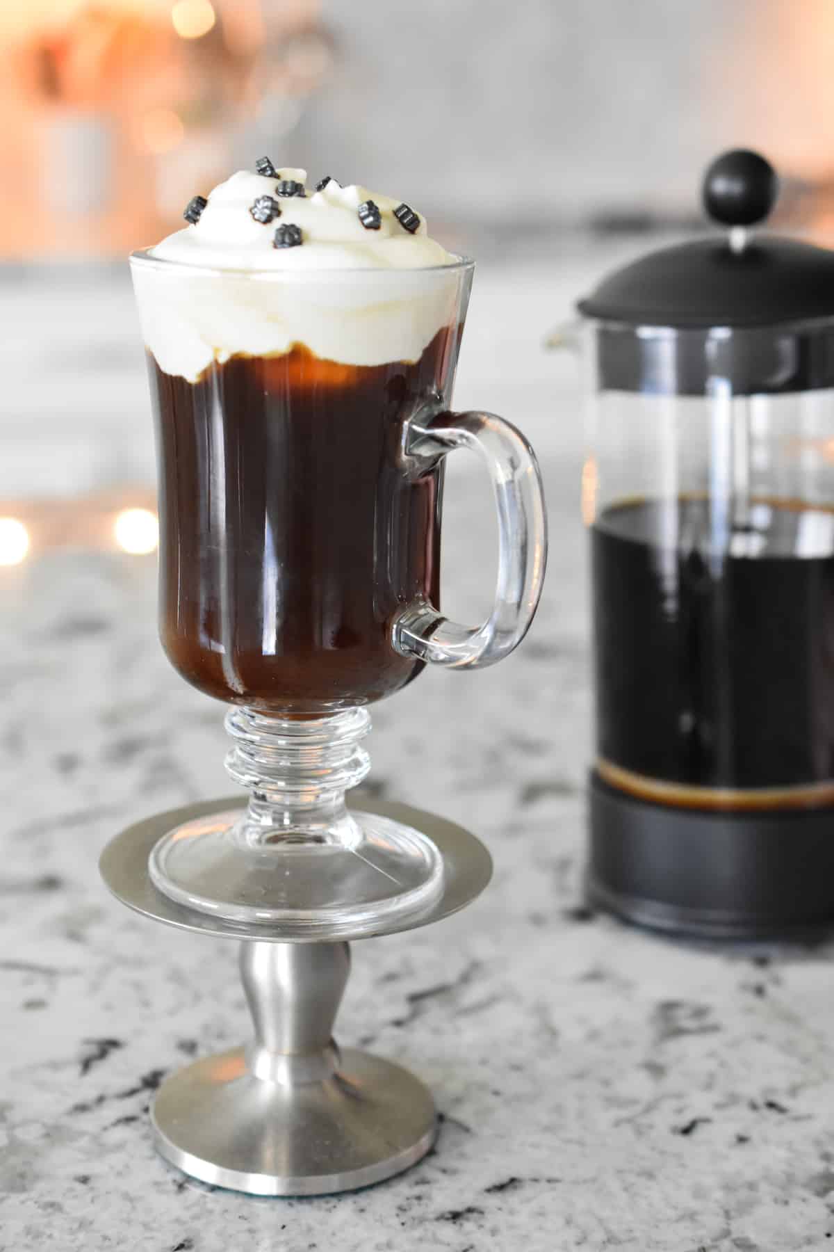 A hot coffee beverage in a footed mug topped with lightly whipped cream. A french press coffee maker is half full with coffee in the background.