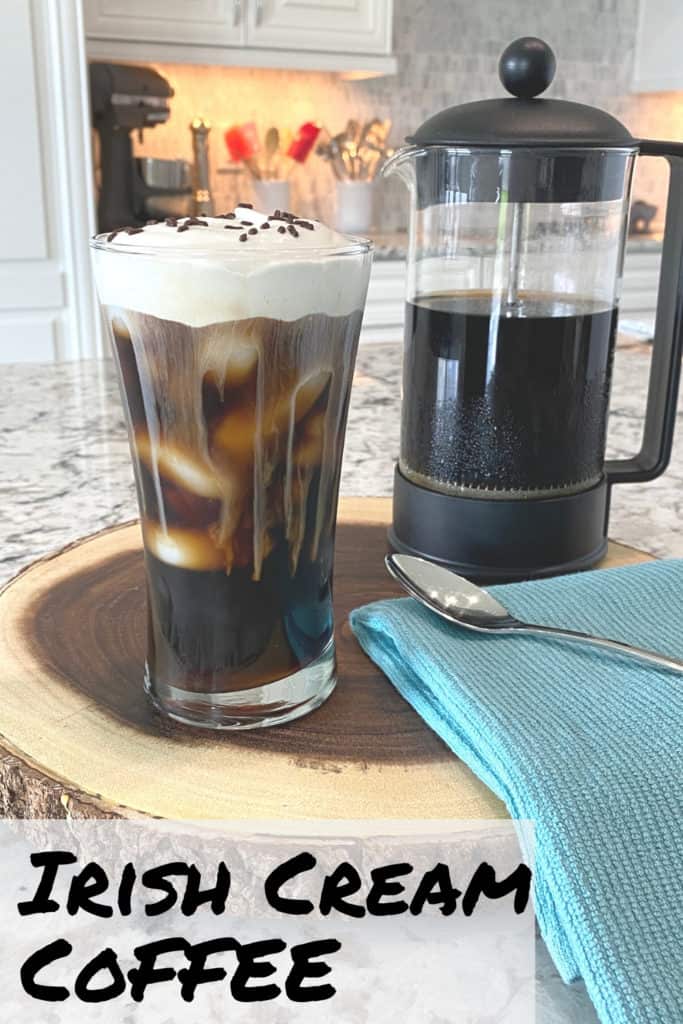 An iced coffee with sweet whipped cream on a cut log tray. A half-filled French press coffee maker is toward the back of the image and a blue towel and spoon is in the foreground.