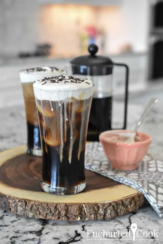 An iced coffee in a tall glass with a creamy topping. A half-filled french press coffee maker is in the background.