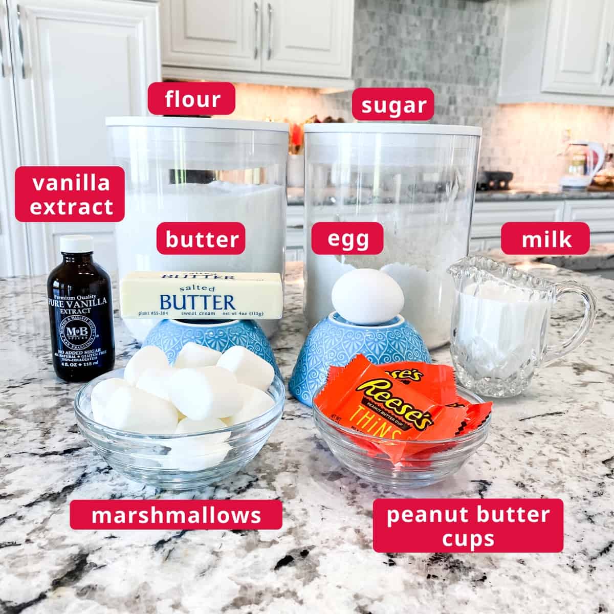 Ingredients for recipe are pictured and labeled. Clockwise from top right: sugar, egg, milk, peanut butter cups, marshmallows, butter, vanilla extract, and flour.