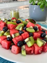 Square image of bright red cubes of watermelon, green kiwi, and blueberries piled on a white platter. A light green dressing is in a carafe along with a potted basil plant is in the background.
