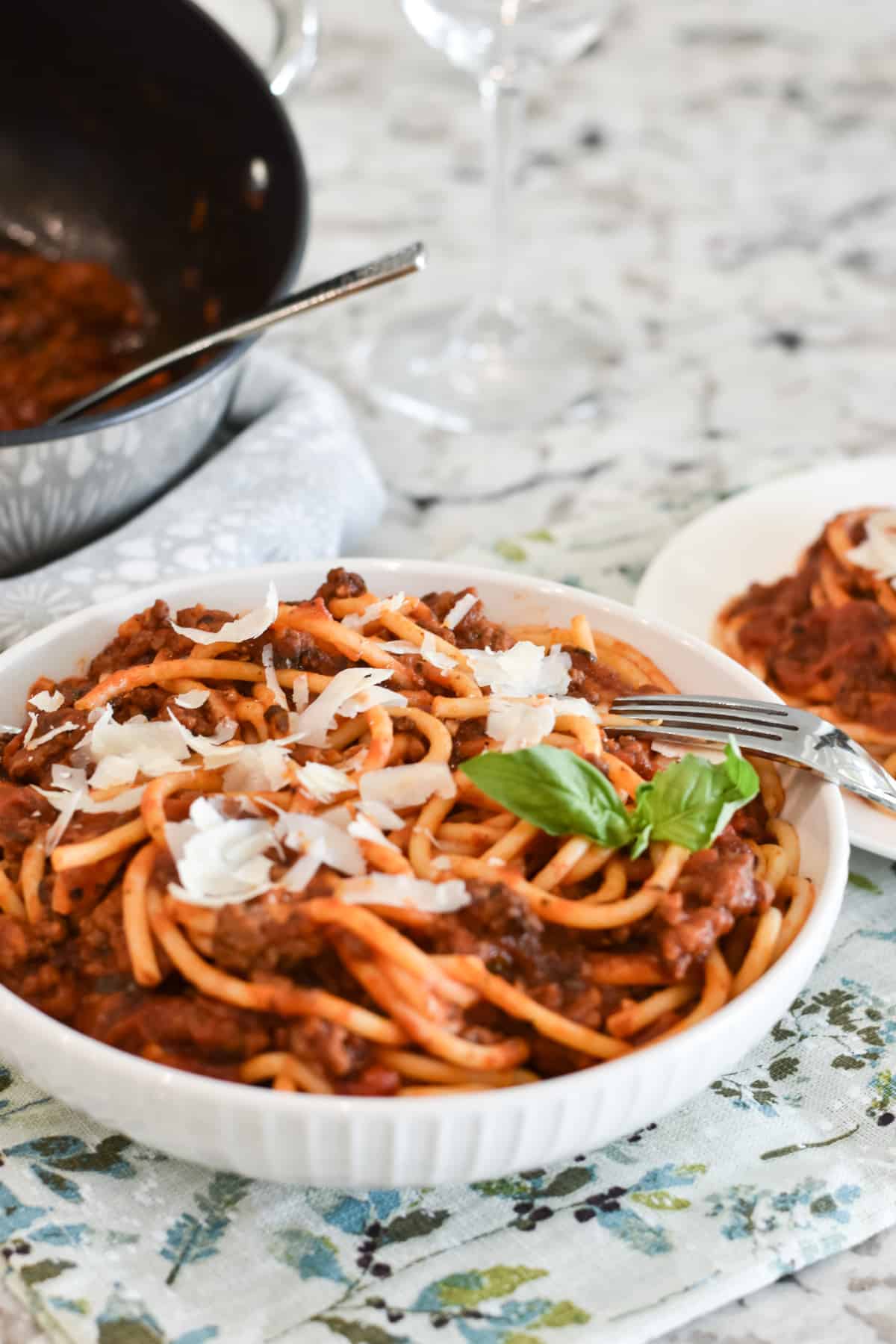 Meat sauce is mixed with bucatini pasta in a white bowl. The pasta is topped with shaved parmesan cheese and garnished with a sprig of fresh basil.