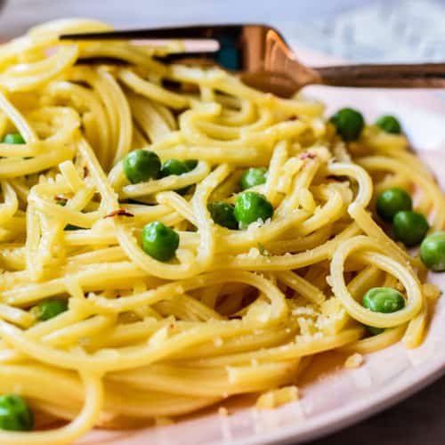 Close up view ot cooked spaghetti with green peas, minced garlic, red peper flakes, and grated parmesan cheese on a pale pink dinner plate.