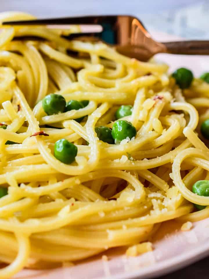 Cooked spaghetti with green peas, minced garlic, red pepper flakes, and grated parmesan cheese on a pale pink dinner plate.