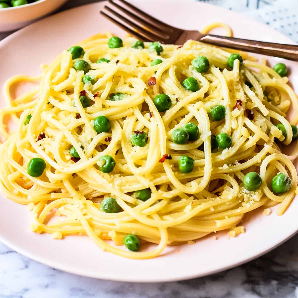 Cooked spaghetti with green peas, minced garlic, red peper flakes, and grated parmesan cheese on a pale pink dinner plate.