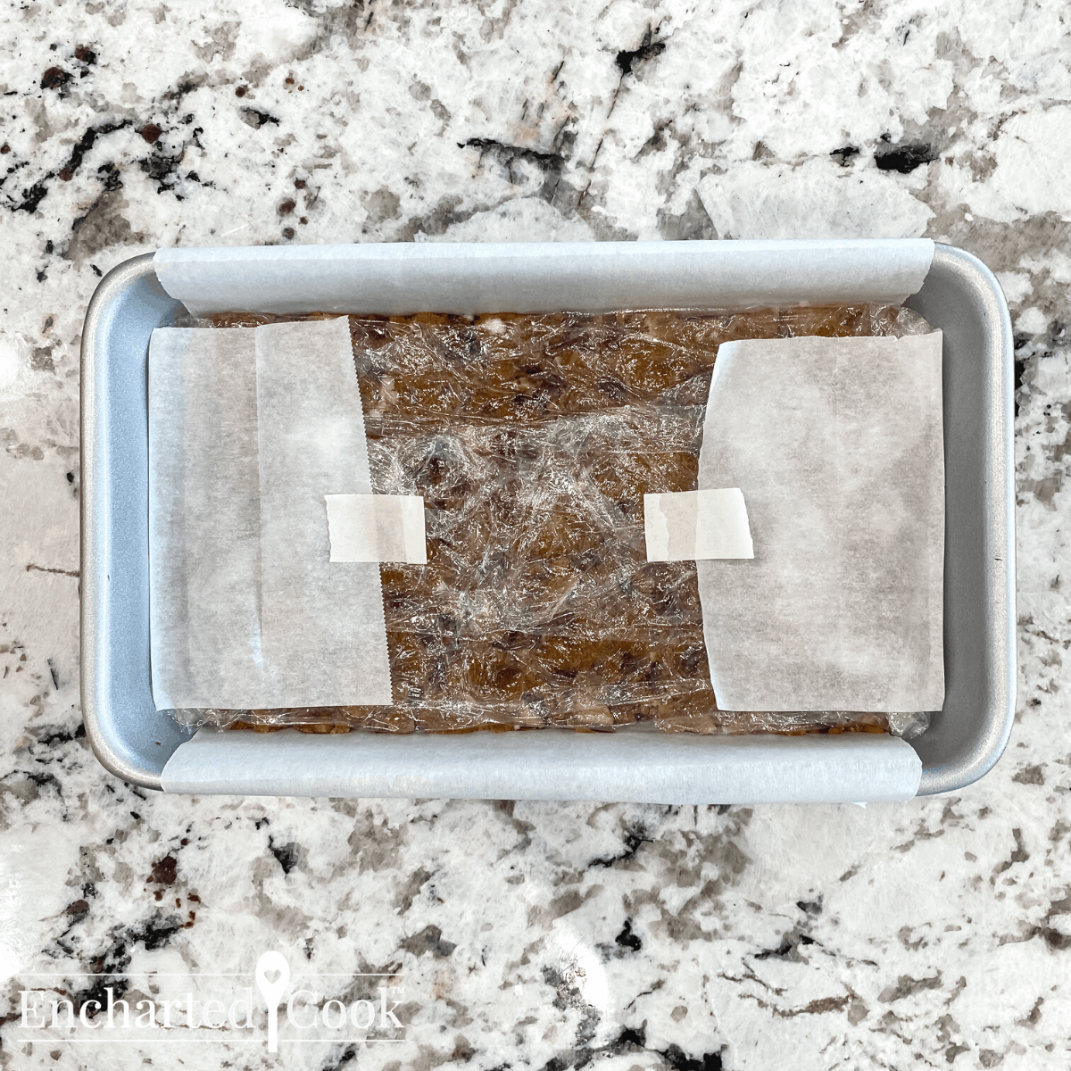 The cake is packed into the parchment paper loaf pan. The top of the fruitcake is covered with plastic wrap.