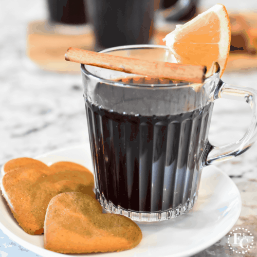 Square image and close up view of deep red wine in a clear glass mug garnished with a slice of orange and a cinnamon stick.