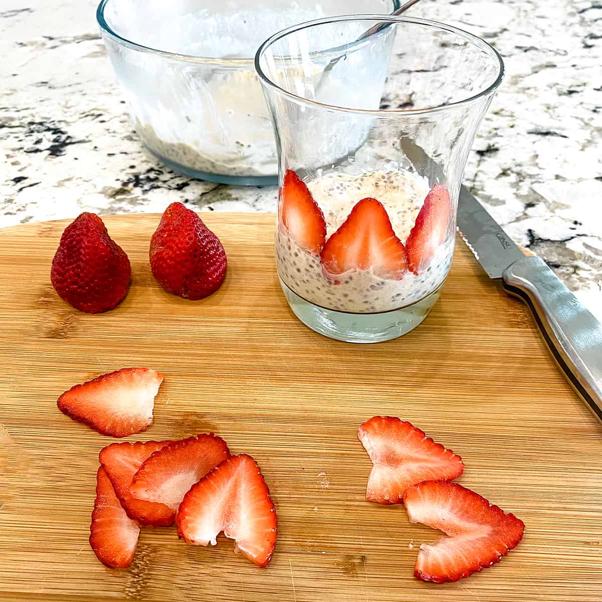 Thinly sliced strawberries are on a cutting board. A clear glass has a layer of oatmeal and slices of strawberries are creating a layer of lovely fruit.