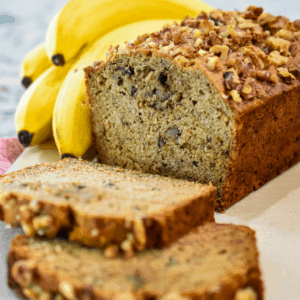 Square image of a sliced loaf of banana bread with bananas in the background.