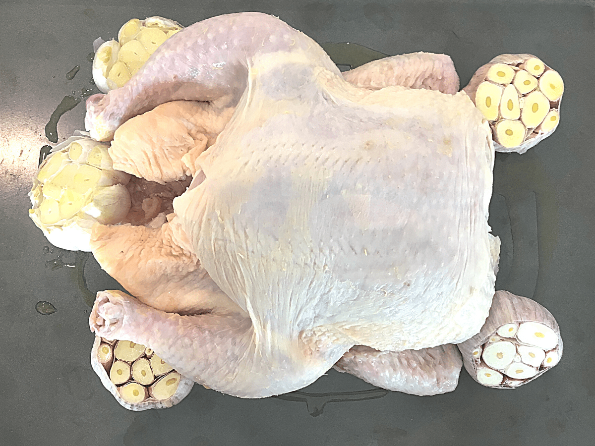 A whole roasting chicken is placed in the middle of the oiled baking sheet and the garlic bulbs are tucked around the chicken.