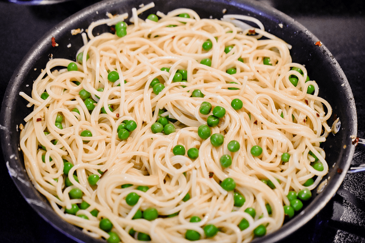 Cooked spaghetti, peas, and the garlic-olive oil sauce is combined in a skillet.