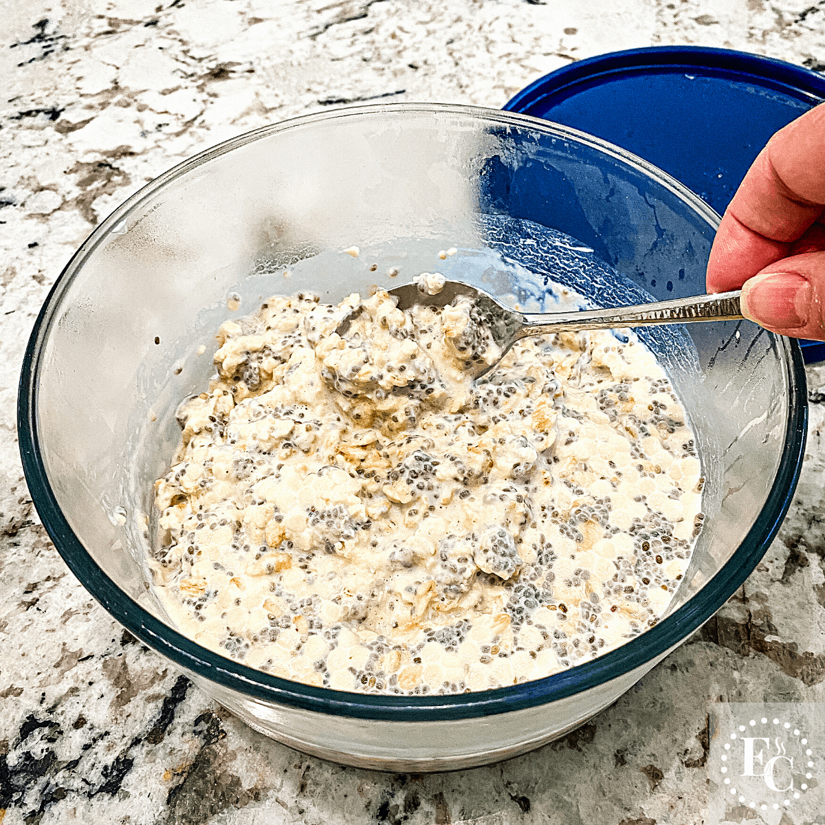 A glass bowl of thickened overnight oats being stirred with a teaspoon.