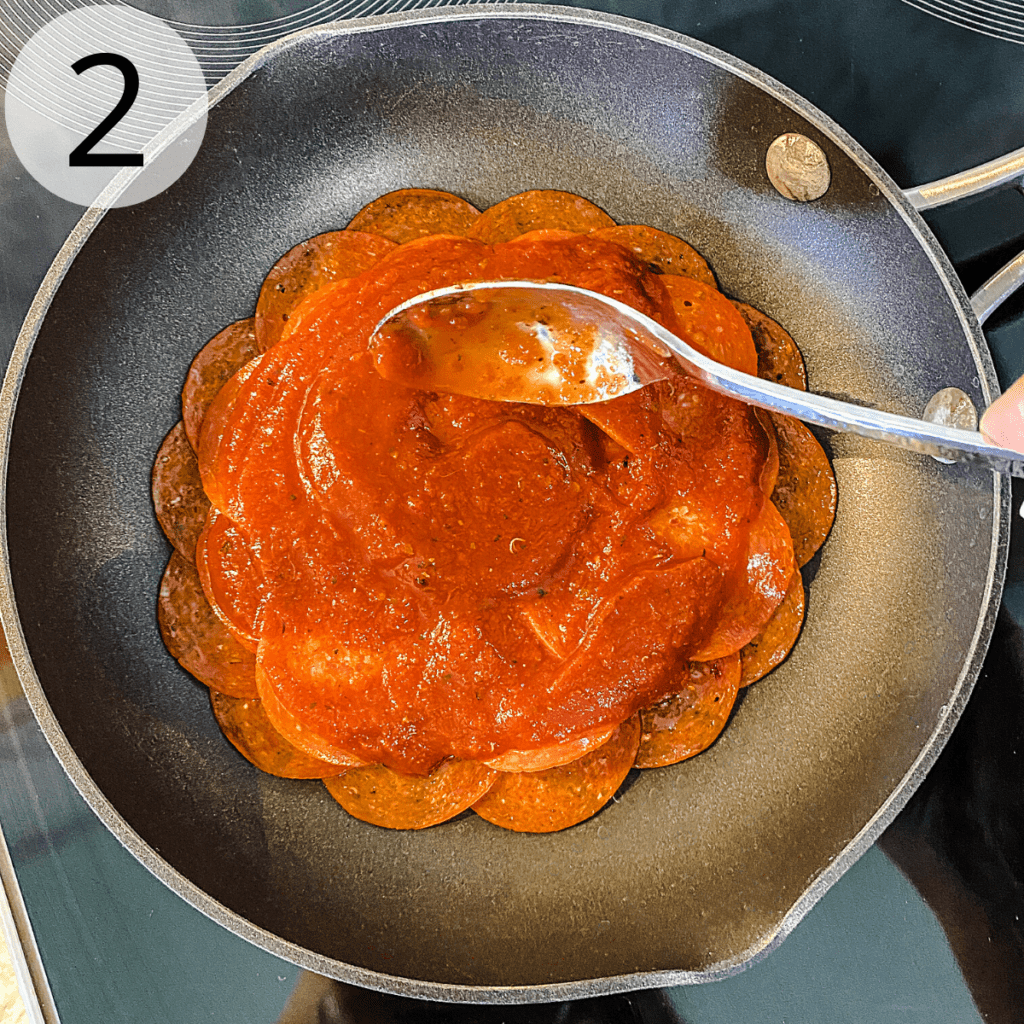 A spoon is being used to add a layer of pizza sauce over the layers of pepperoni in a small fry pan.