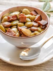 Lima beans and pieces of pink ham in a small bowl with a plate and spoon.