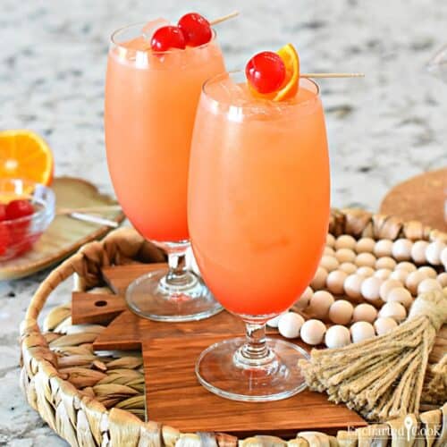 Two tall fruity cocktails garnished with a slice of orange and maraschino cherries.