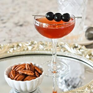 The cocktail in a coupe glass garnished with cocktail cherries.