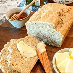 Square image of Beer Bread sliced on a dark cutting board with honey and slices of butter on the side.