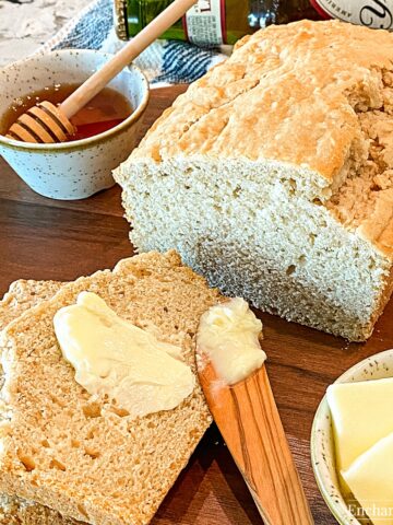 Square image of Beer Bread sliced on a dark cutting board with honey and slices of butter on the side.