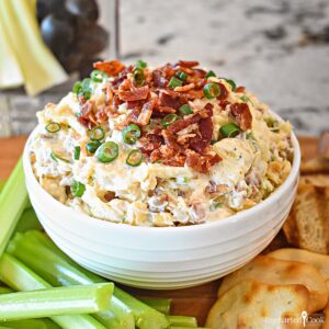 Square image of dip in a white bowl topped with bacon and green onion surrounded by celery sticks and crackers.