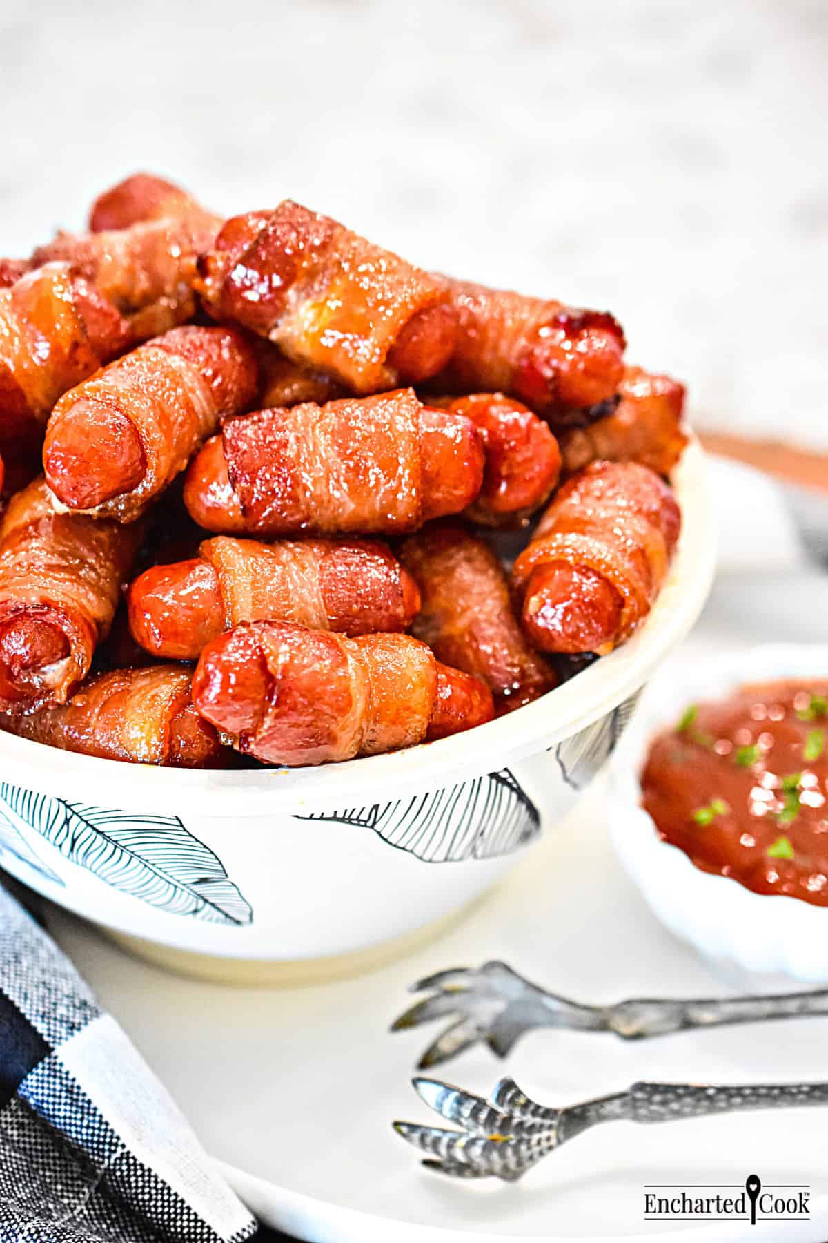 Bacon Wrapped Smokies in a bowl with a black and white napkin and decorative tongs.