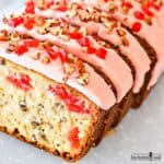 A sliced cherry pecan loaf cake frosted with pink frosting and decorated with chopped pecans and cherries on a white plate.