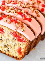 A sliced cherry pecan loaf cake frosted with pink frosting and decorated with chopped pecans and cherries on a white plate.