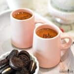 Creamy Hot Chocolate is in two small pink mugs and garnished with chocolate sprinkles. A small dish of mini oreos is in the foreground.