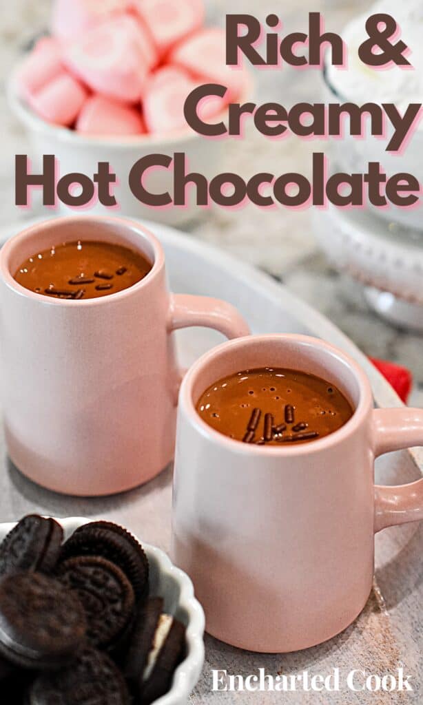 Creamy Hot Chocolate is in two small pink mugs and garnished with chocolate sprinkles with text overlay.