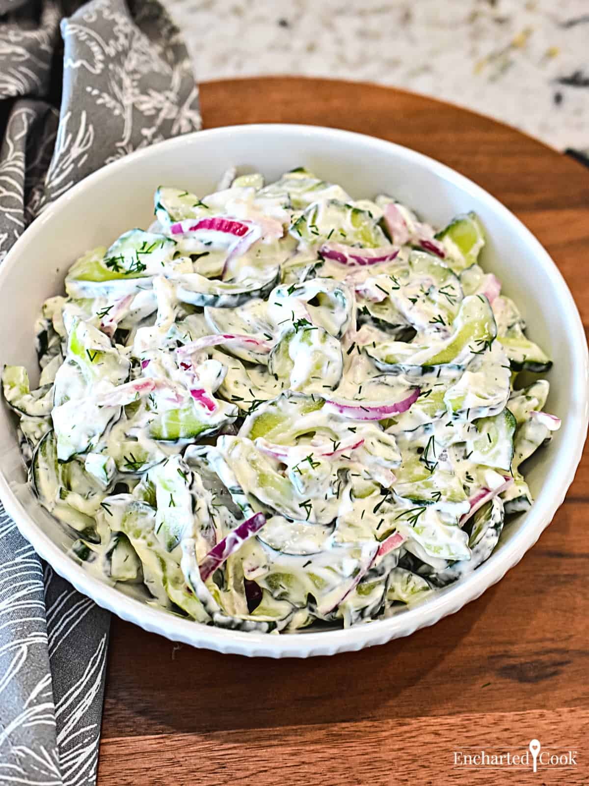 Sliced cucumber and red onion in a creamy dressing in a white bowl on a wooden board.