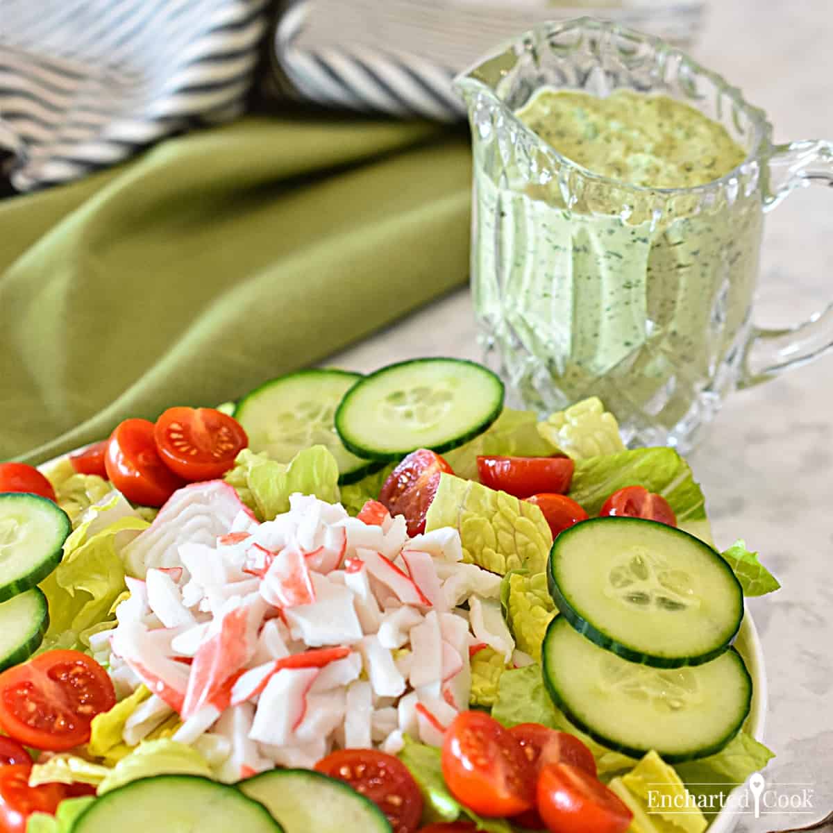 A salad of lettuce, tomato, cucumber, and krab, with green salad dressing in a crystal pitcher.