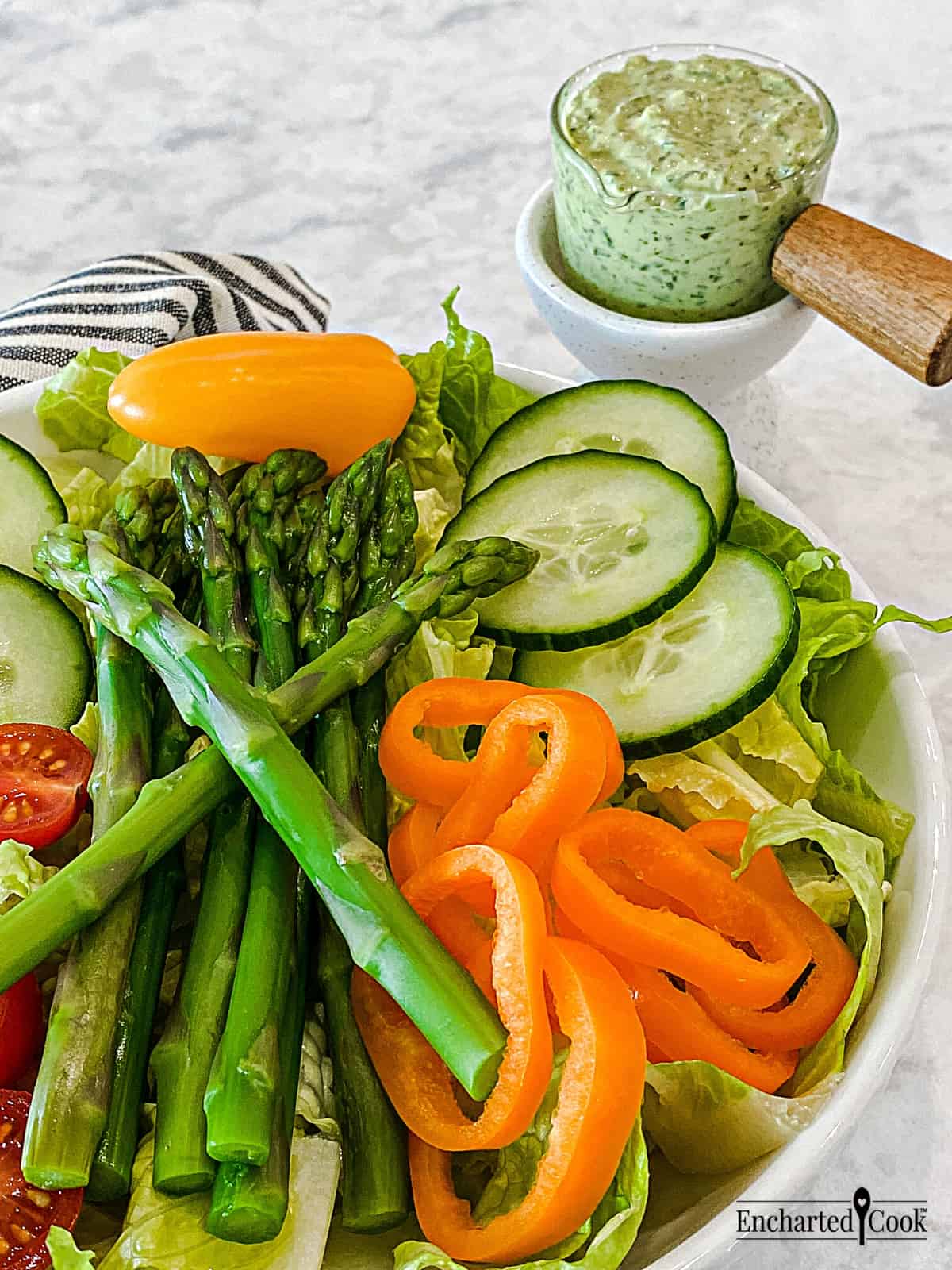 A garden salad of lettuce, cucumber, asparagus, orange peppers, and cherry tomatoes, with green salad dressing in a small pitcher.
