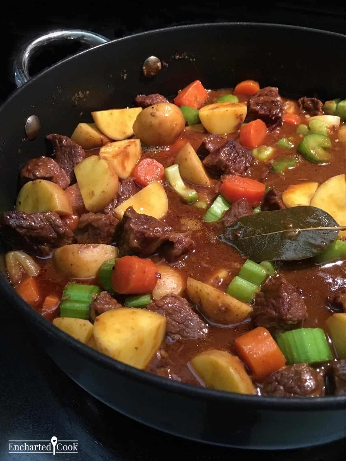 Beef, potatoes, carrots, and celery in a rich brown gravy with a bay leaf in a large pan.