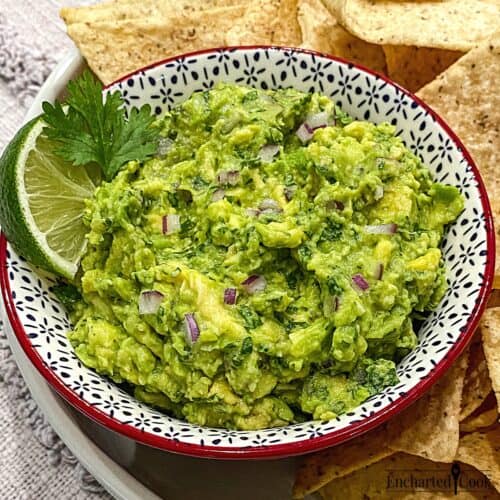 A bowl of freshly made green guacamole garnished with lime and cilantro with tortilla chips surrounding it.