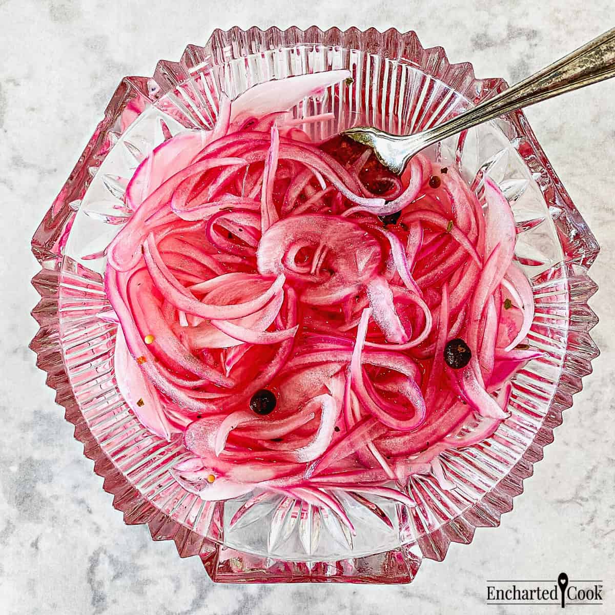 Slices of pink-colored pickled red onion in a fancy-cut glass dish with a fork.