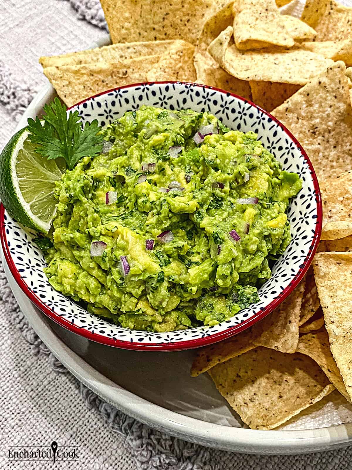 A bowl of freshly made green guacamole garnished with lime and cilantro with tortilla chips surrounding it.