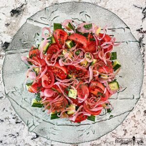 Wedges of bright red tomatoes are mixed with cucumber and pickled red onions in a large green glass bowl.
