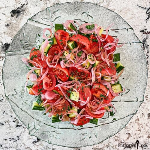 Wedges of bright red tomatoes are mixed with cucumber and pickled red onions in a large green glass bowl.
