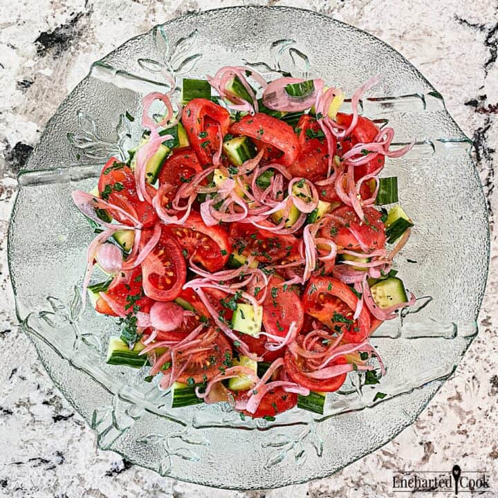 Wedges of bright red tomatoes are mixed with cucumber and pickled red onions in a large green glass bowl.