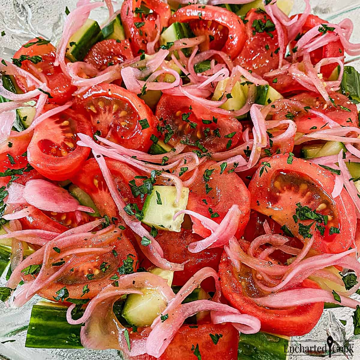 Close up view of wedges of bright red tomatoes are mixed with cucumber and pickled red onions in a large green glass bowl.